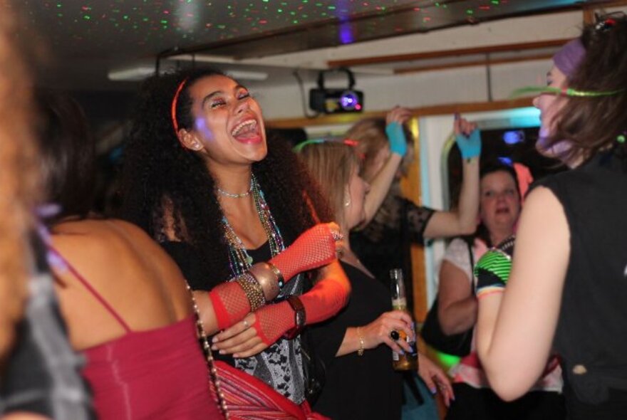 People dancing on board a boat, dressed in 1980s fashions including gloves headbands and jewellery.