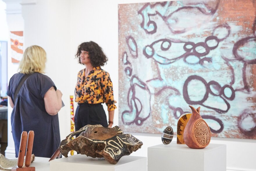 People in a gallery talking to each other, with a large artwork on the wall, and small sculptures on white display plinths.