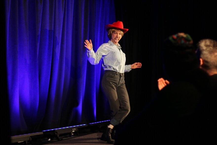 A comedian on a stage wearing a red cowboy hat, performing in front of a live audience.