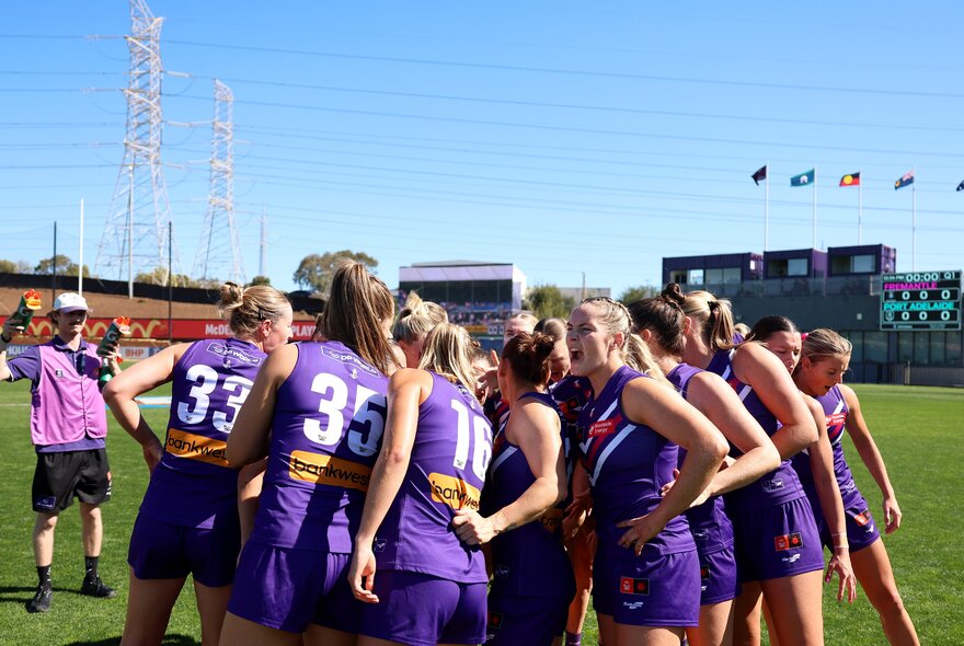 Fremantle AFLW players celebrate in a huddle on a bright sunny day. 