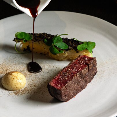 Sauce being poured from above onto a white dining plate over a tenderloin of beef, with a vegetable side.