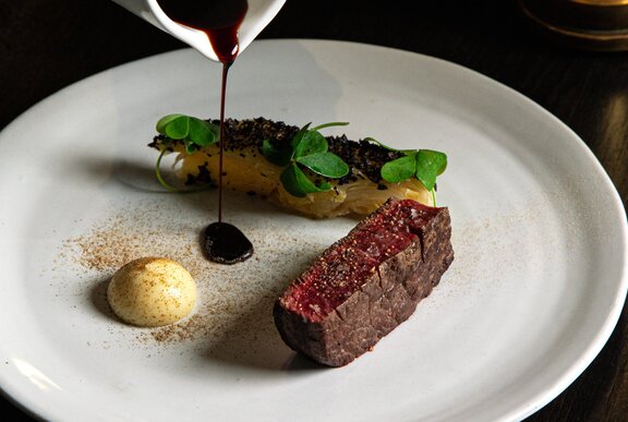 Sauce being poured from above onto a white dining plate over a tenderloin of beef, with a vegetable side.