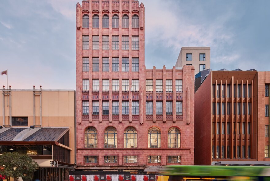 A tram going past a multi storey pink building in the city.