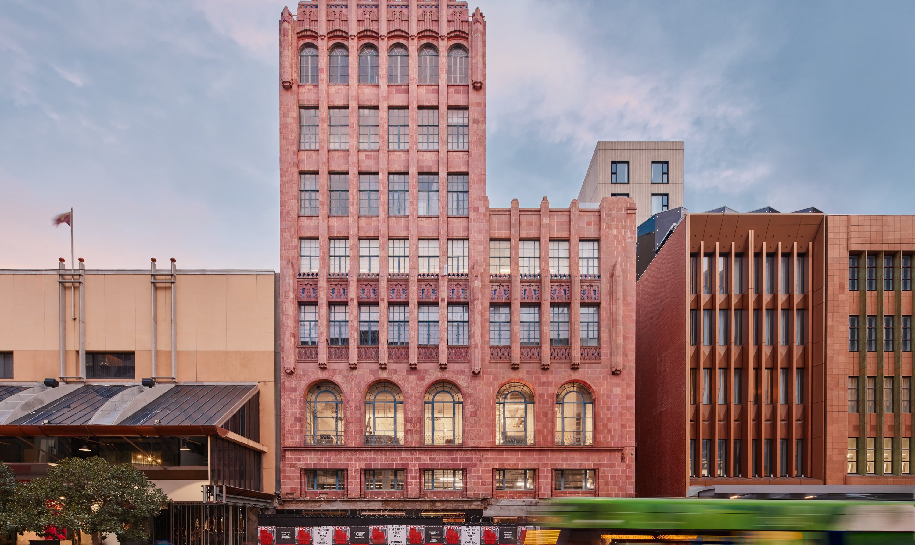 A tram going past a multi storey pink building in the city.