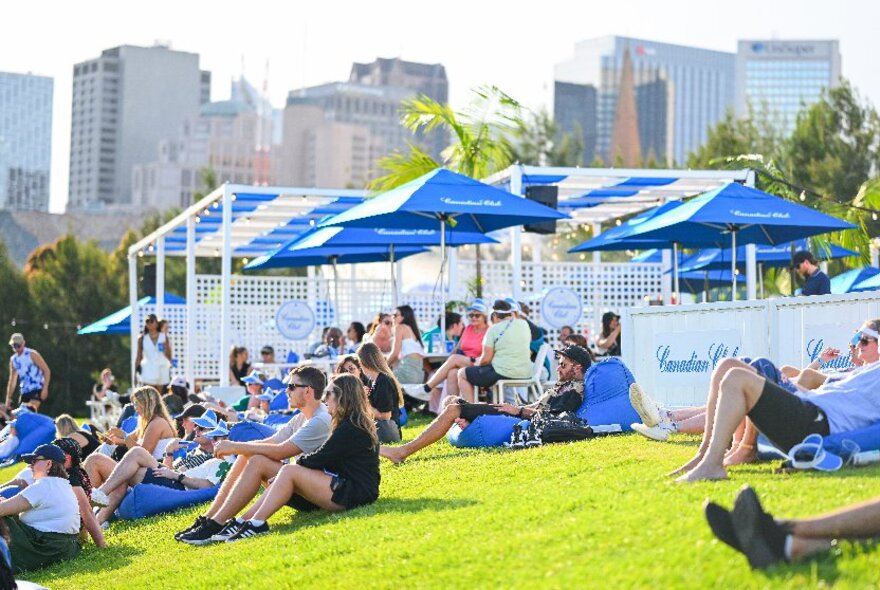 People seated on a grassy sloped area of Birrarung Marr park, home to AO Hilltop, an area with some seats, food and drink stalls and blue shade umbrellas. 