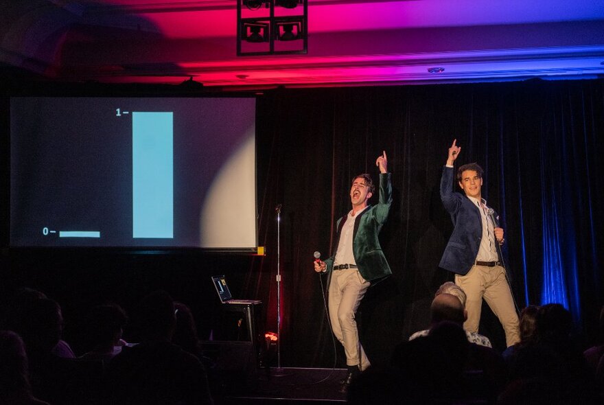 Two people performing on a small stage in front of a seated audience, a large screen to their right, both of them pointing an arm high into the air.