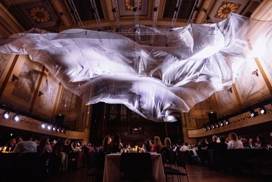 The interior of the Royal Exhibition Building with a gauzy white sheet hanging from the ceiling, patrons seated at long tables for dinner.