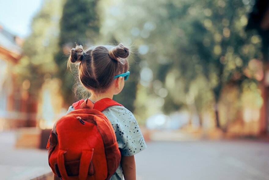 The back view of a young child wearing a small school rucksack on their back, their hair braided into two buns on either side of their head, walking outside.