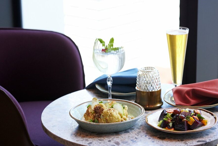 Dark purple chair to left of table with two dishes of food, one cocktail adenine long flute at rear.