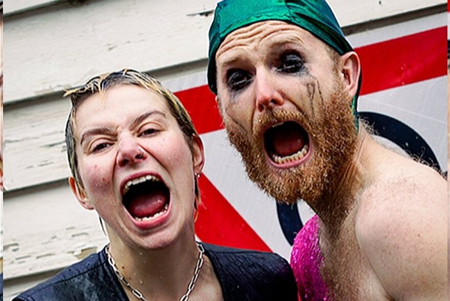 Two performers outside, both screaming with open mouths, one of them has smudged eye make up, a beard and is wearing a green swimming cap on his head.