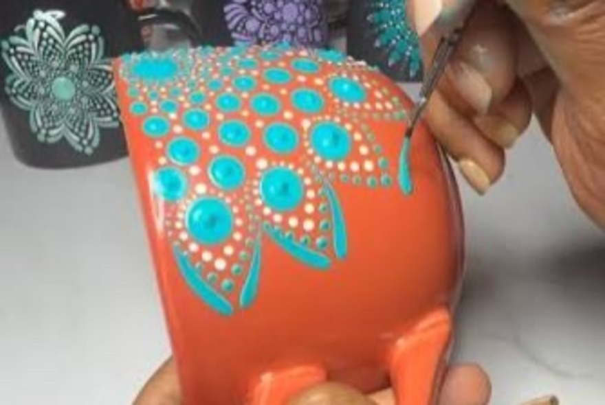 A person using a small brush to create a dot pattern design with turquoise and white acrylic paint on an orange ceramic mug, as part of a creative workshop.