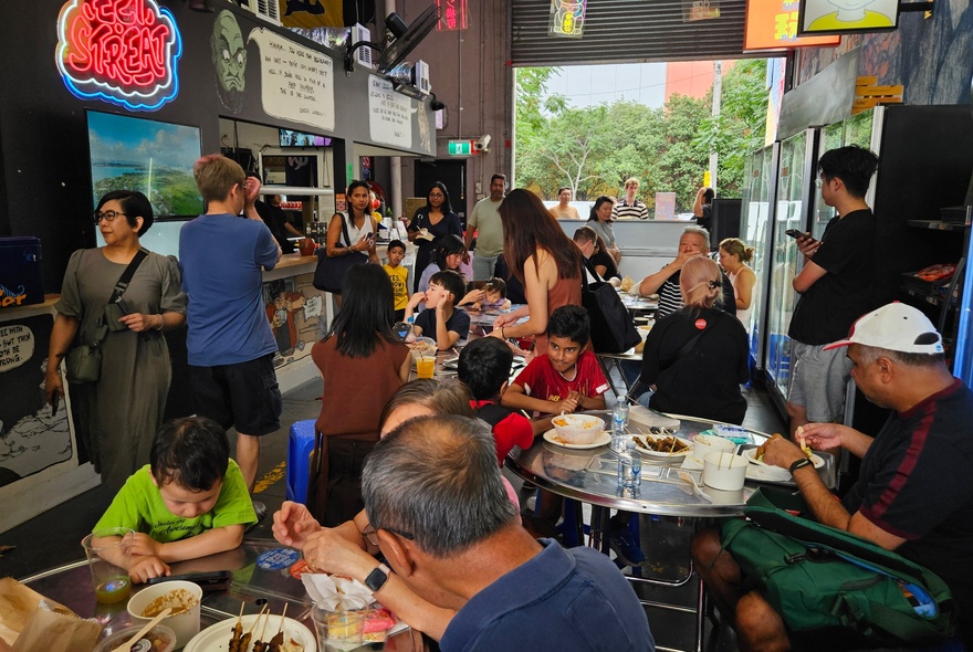 People seated at tables in a food hall, dining on a range of food and drink.