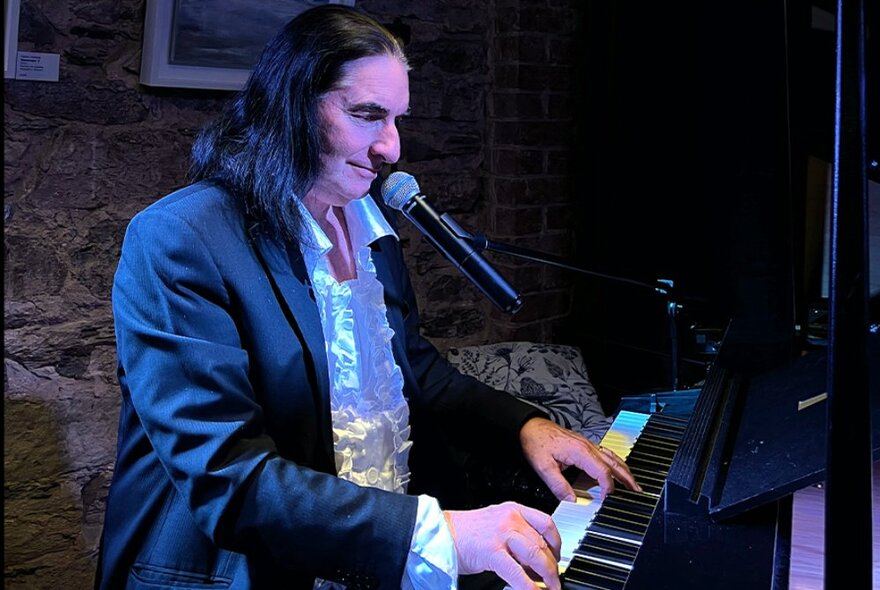 A performer dressed as Meat Loaf in a suit jacket and ruffled shirt, seated at a grand piano and singing into a microphone.