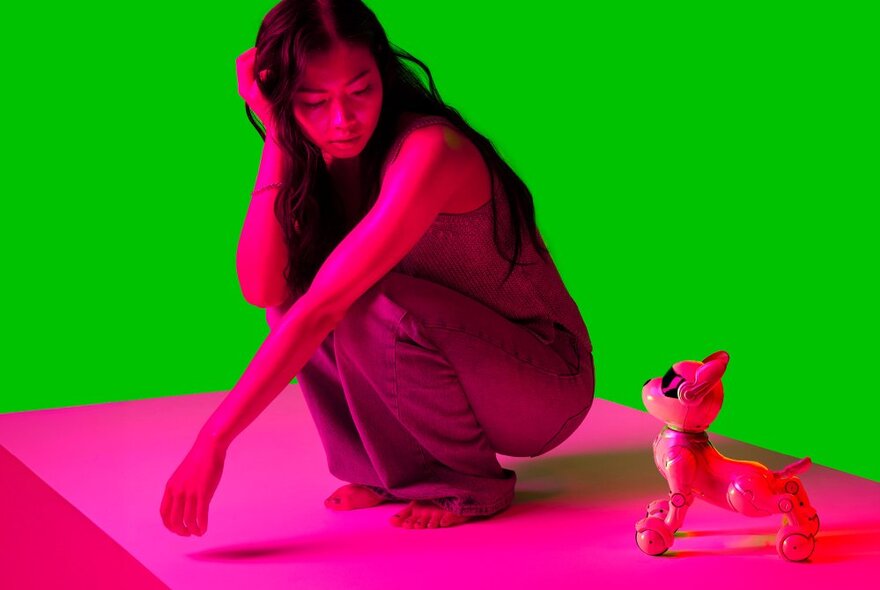 Bright green and pink image of a girl squatting on a platform next to a robot dog toy.