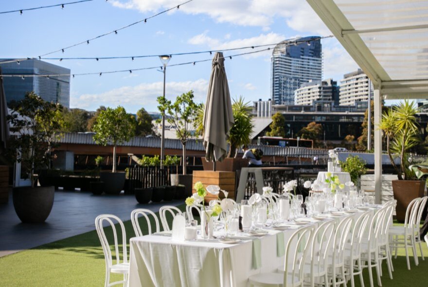 Long table set for dining on an outdoor terrace with city buildings in the background.