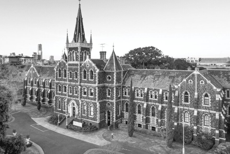 A black and white picture of Deaf Children Australia's historic Bluestone building with a pointy tower in a large garden setting. 