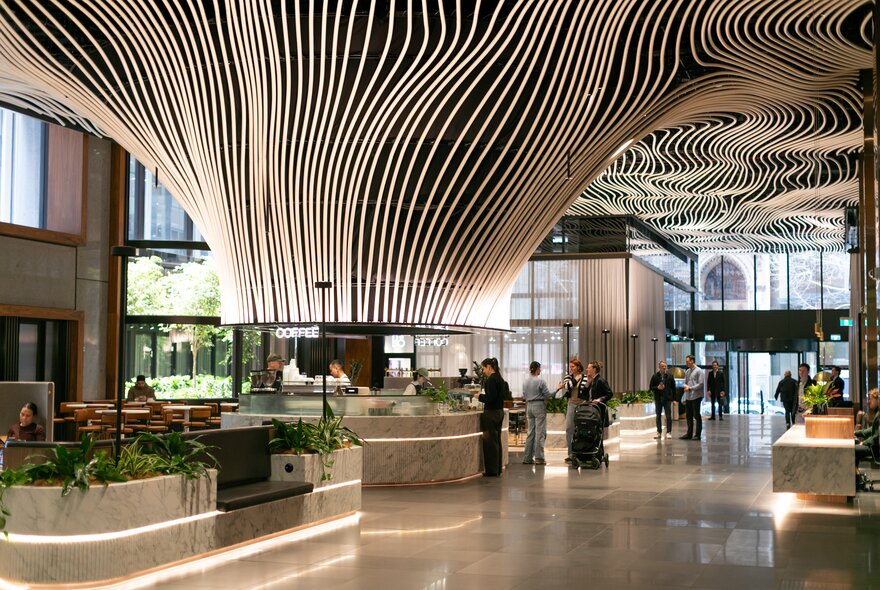 Foyer of a city building with an indoor architectural space featuring a central modern coffee bar with a striking ceiling installation made of numerous curved, flowing white lines that converge above the counter.