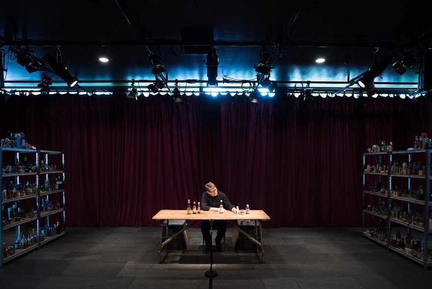 A man in black seated at a wooden table on stage, carefully positioning condiment bottles on the table.
