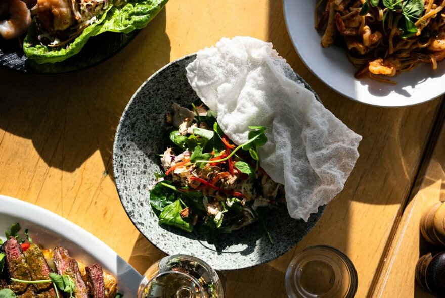 Plates of food arranged on a table including a central bowl of salad with crispy thin flatbread resting on the edge of the bowl.