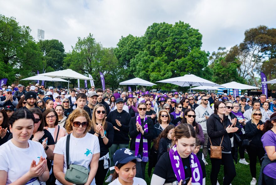 Large crowd gathered in a park with umbrellas, wearing purple scarves.