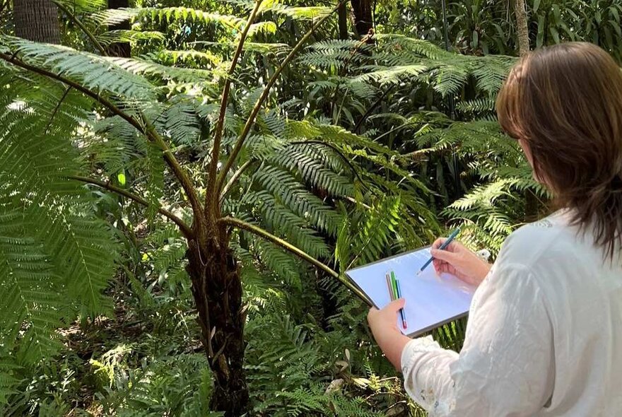 A person standing outside in front of fern plants, drawing onto a white sketch pad that she holds in her hand. 