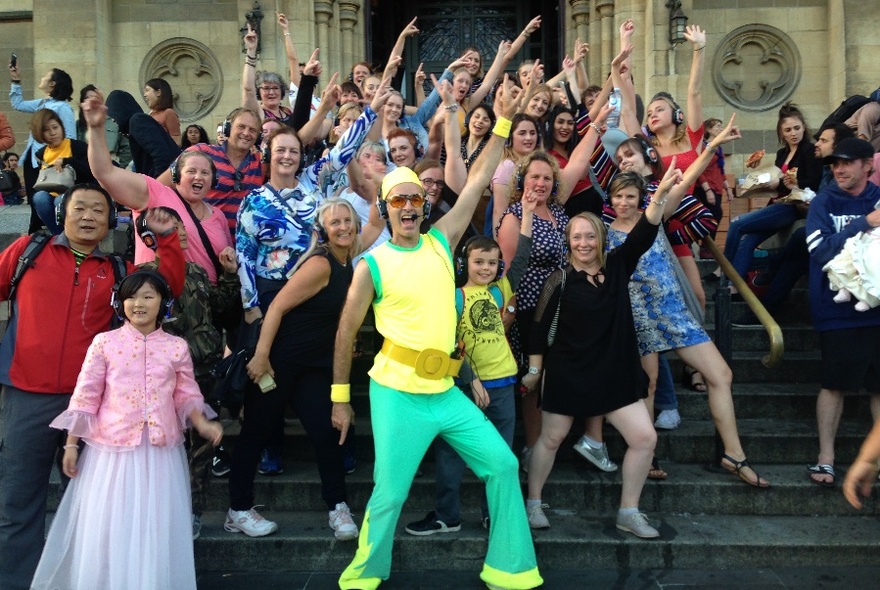 Group dancing on stairs of a sandstone building, led by man in bright green and yellow shiny outfit, all wearing headphones.