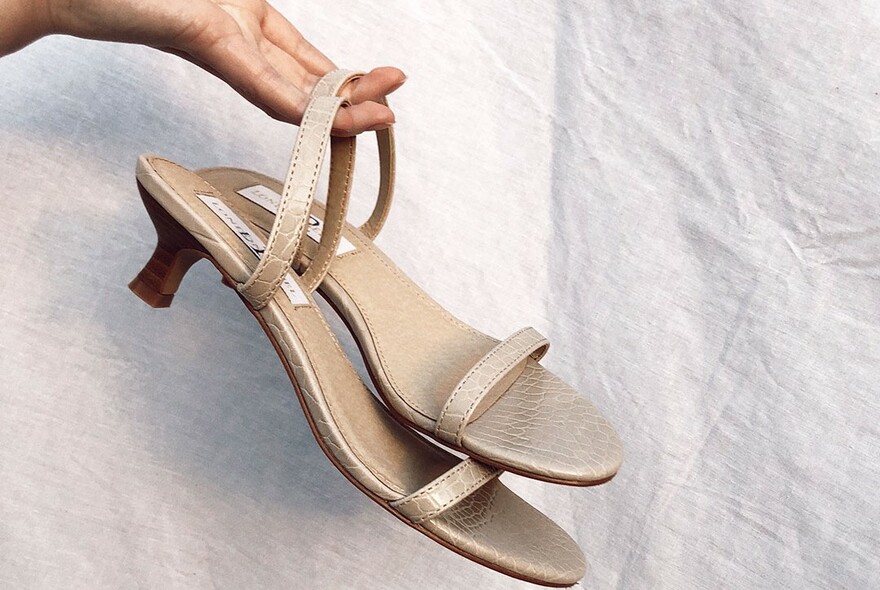 A hand holding a pair of kitten-heel beige sandals against a linen curtain.