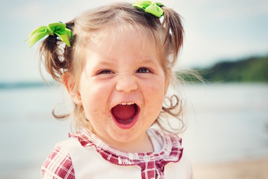 A young child laughing with an open mouth, their hair styled in high short pigtails tied with green ribbon, a beach backdrop.