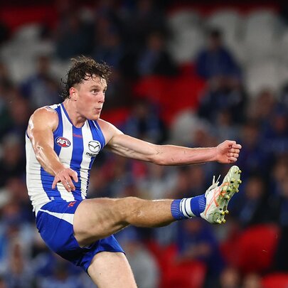 A North Melbourne AFL player with his leg raised high in front of him, having just kicked a football, with blurred fans in the stadium in the background.