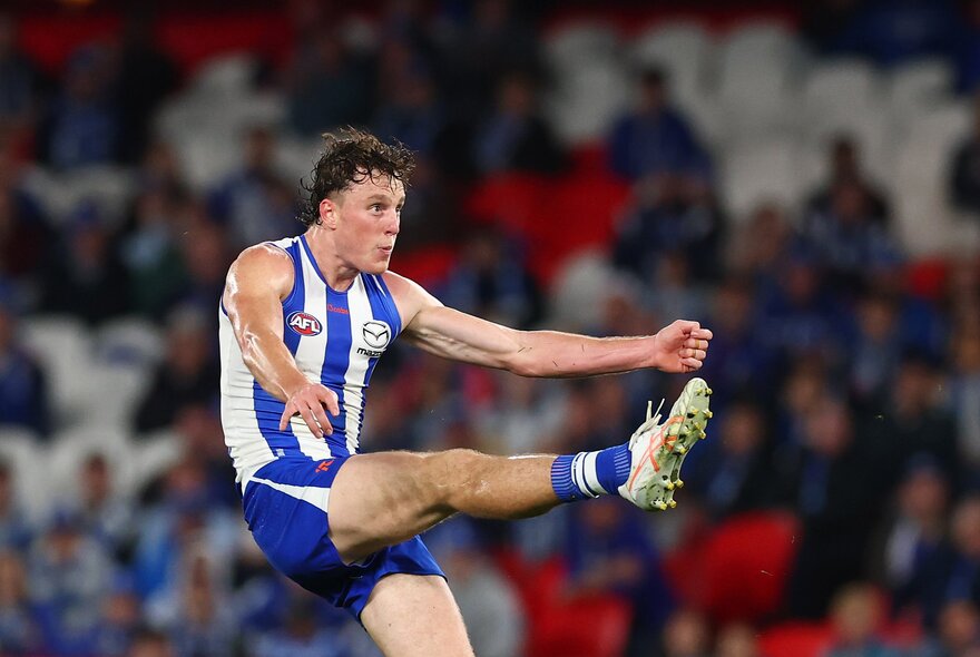 A North Melbourne AFL player with his leg raised high in front of him, having just kicked a football, with blurred fans in the stadium in the background.