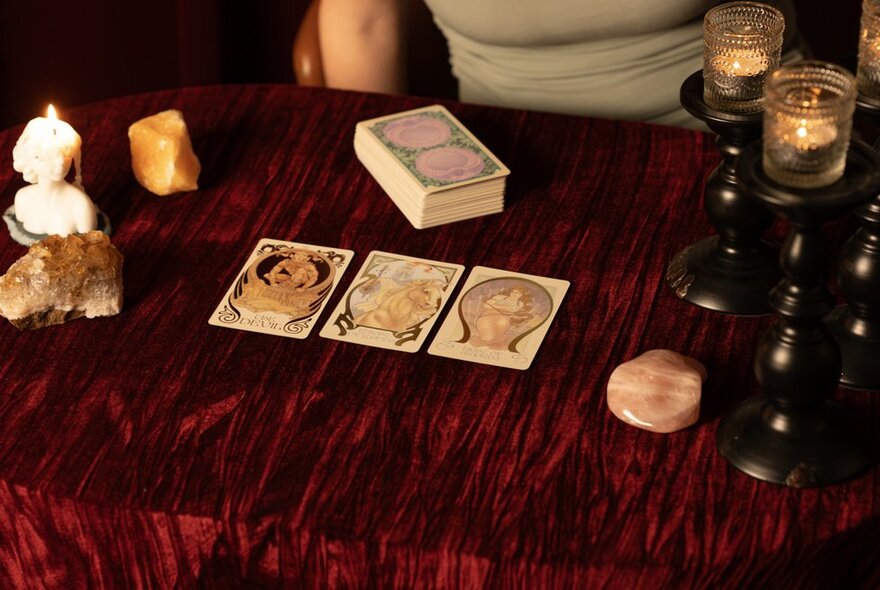 A person performing a tarot card reading on a velvet-draped table with three face-up cards, a deck stack, candles, and crystals.