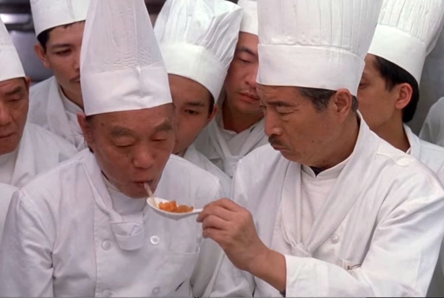 A group of chefs, all wearing white uniforms and chef hats, one of them tasting food from a spoon that another chef holds; a still from the film, Eat Drink Man Woman.