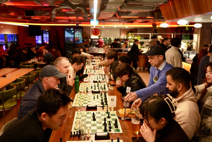 Rows of chess players with boards and chess pieces set up on a long table in an indoor venue.