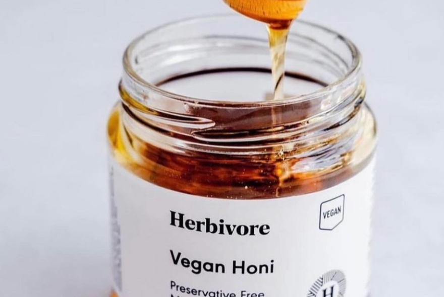 A wooden spoon lifting up honey from a glass jar with the words 'Vegan Honi' written on a  white label on the jar.