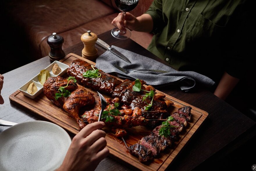 Two people seated opposite each other at a restaurant dining table, one person's hand using a fork to reach into a platter of grilled meats and poultry.