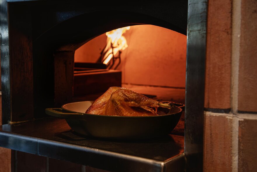 A whole chicken in a baking dish in the doorway of a wood-fired oven, flames visible inside. 