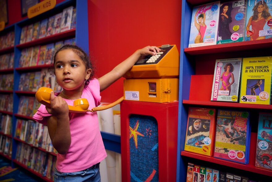 A young child playing with an old orange Telecom pay phone in an exhibition space designed to look like a DVD library.