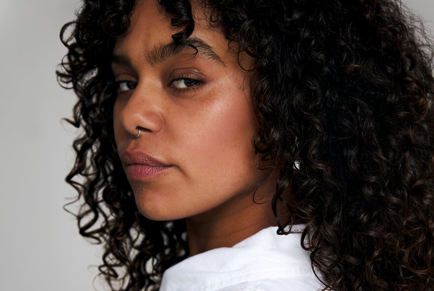 Woman with curly black hair, her head turned and looking over her shoulder with a neutral expression.