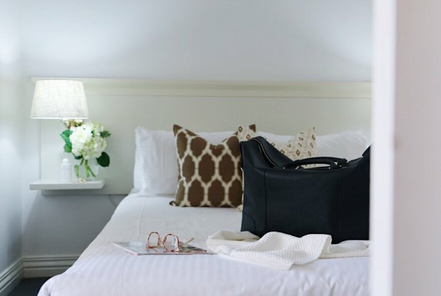 A hotel room bed with white sheets, brown cushions and a black weekender bag sitting on top.