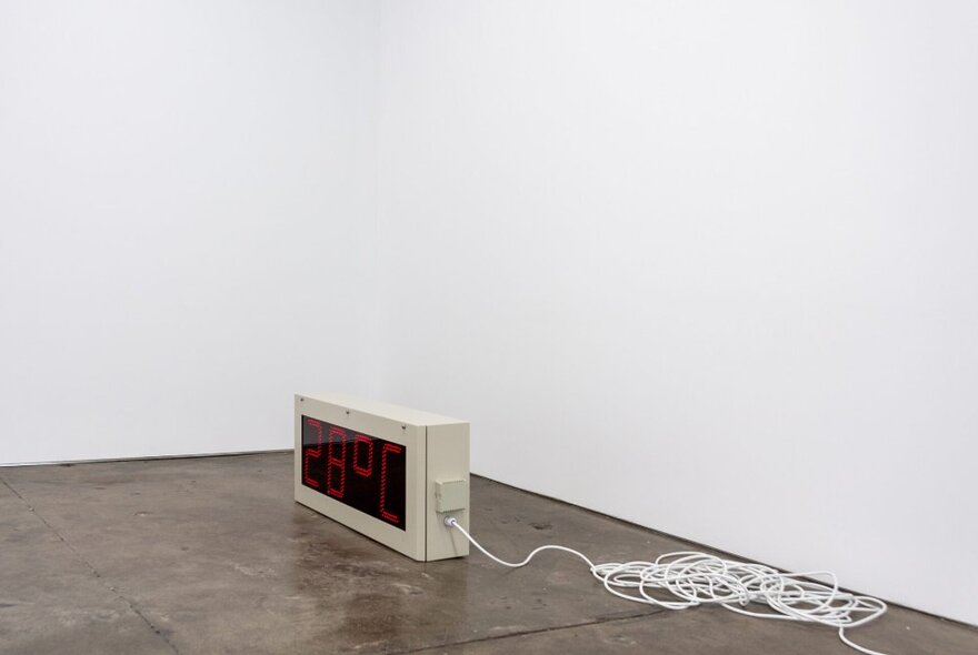 Art installation of grey box connected to a white cable on a shiny brown floor against white walls.