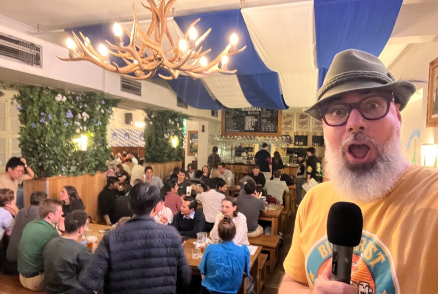 A surprised looking man wearing a Tyrolean hat and holding a microphone in a large room with striped drapes on the ceiling and people seated at benches.