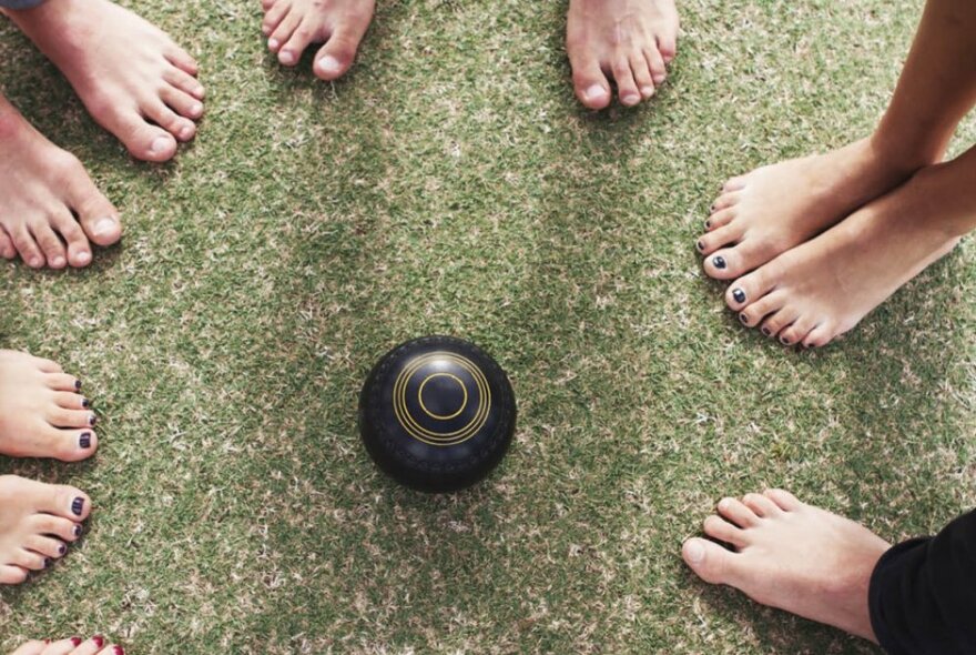 A collection of bare feet gathered around a central lawn bowl on green grass.