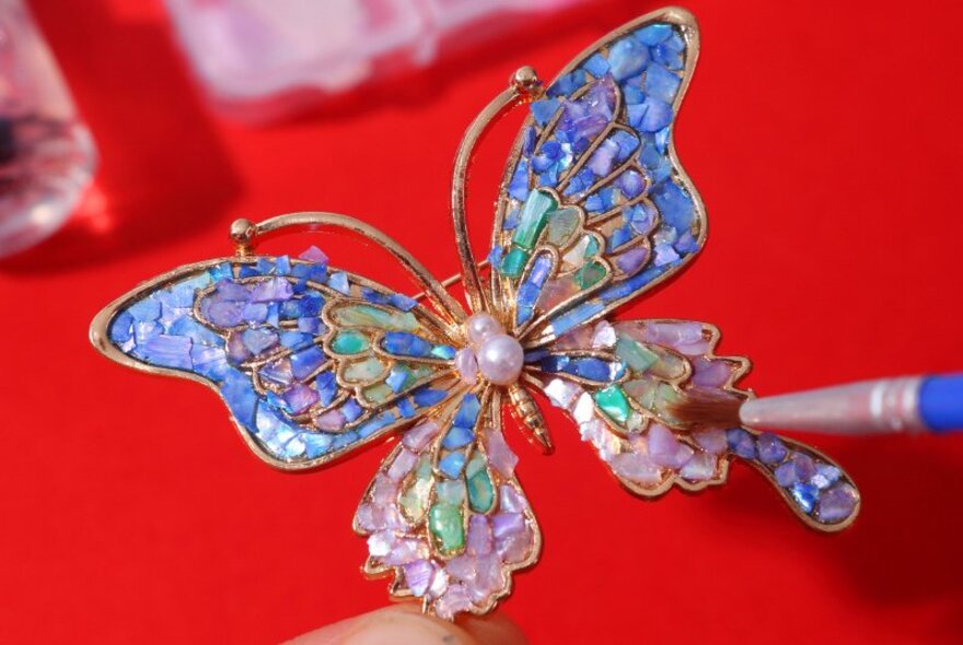 A person using a small brush to seal the mother of pearl inlay on a decorative butterfly brooch, as part of a jewellery workshop.