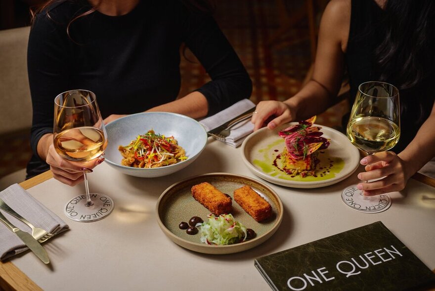 Two diners seated at a restaurant table, plates of food and glasses of wine in front of them.