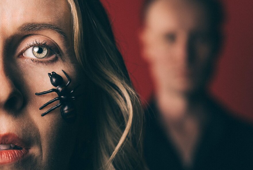 A close-up photograph of a woman's face with a black spider prop near her right eye, and a blurred person in the background.