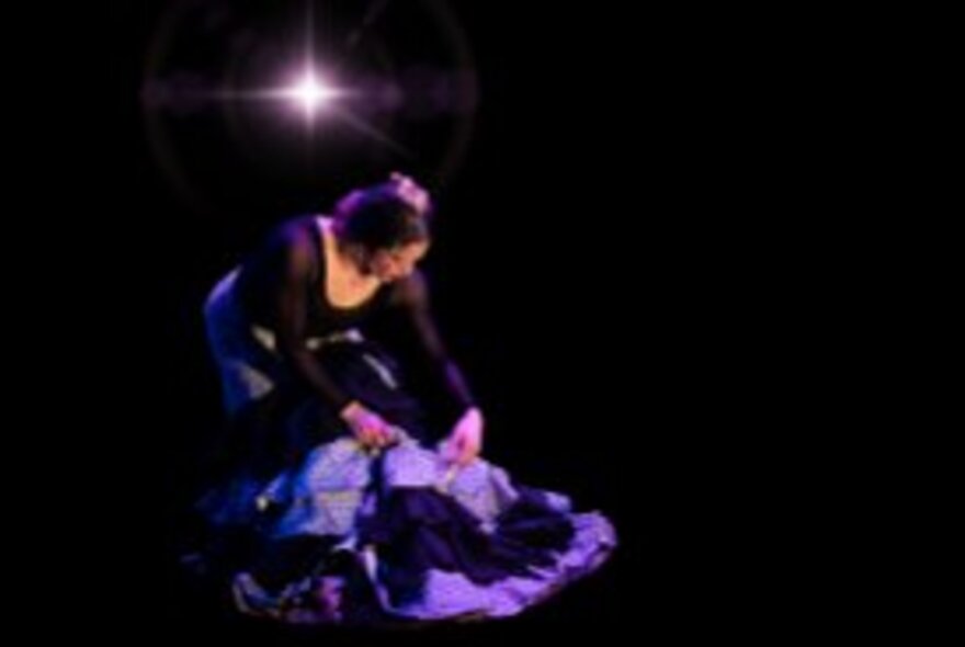 A flamenco dancer on a darkened stage, bending over with her arms almost touching the ground.
