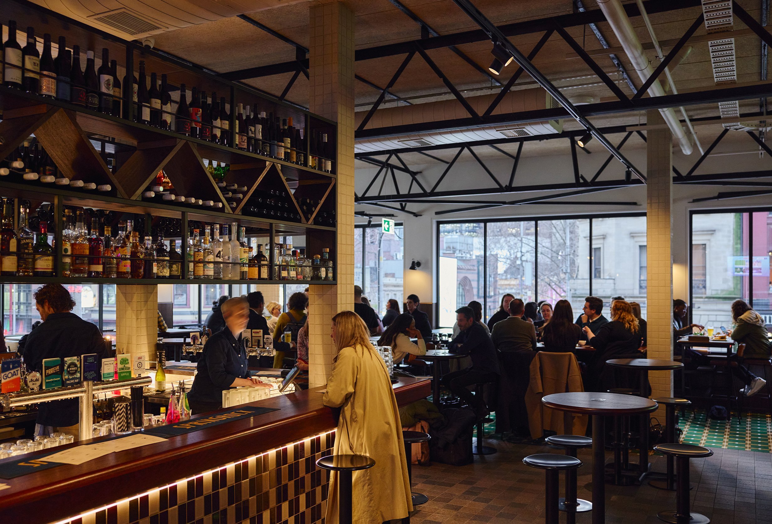 Inside a busy pub people are drinking and a woman is ordering a drink at the bar
