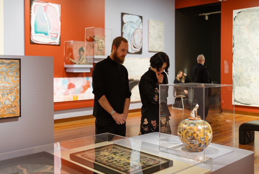 Two people in black in a gallery space looking at artefacts under glass cases.