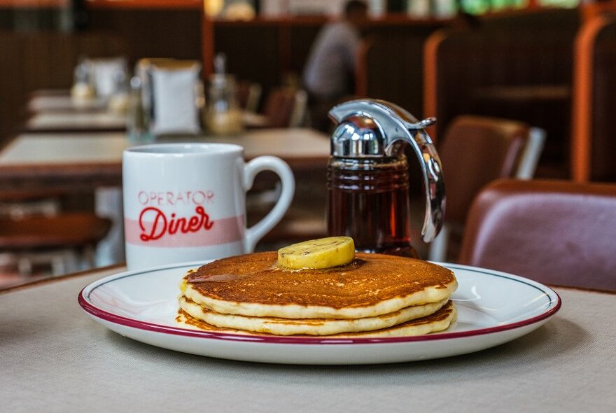 A stack of pancakes with a jug of maple syrup and a mug.