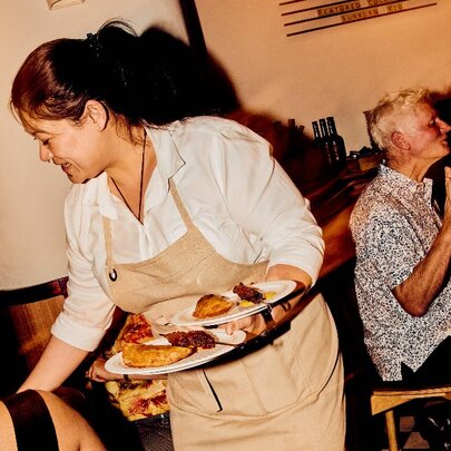A Peruvian waitress wearing a white shirt and beige apron at Barra Restaurant, serving plates of Peruvian food to diners.